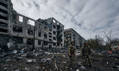 Two soldiers walk past burnt-out blocks of flats with rubble strewn around them.