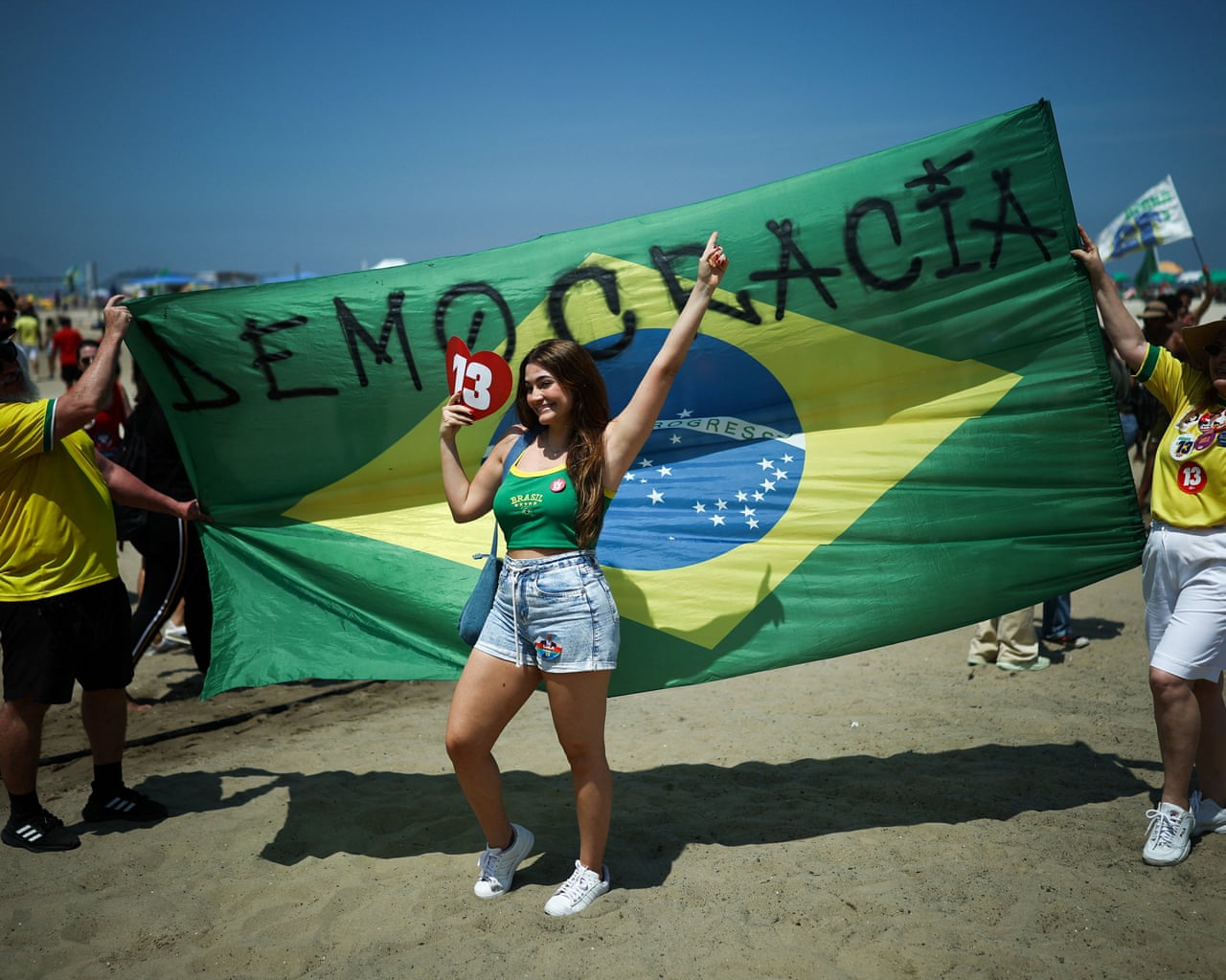 A smiling woman poses before a huge Brazilian flag reading “Democracia” at a Lula rally.