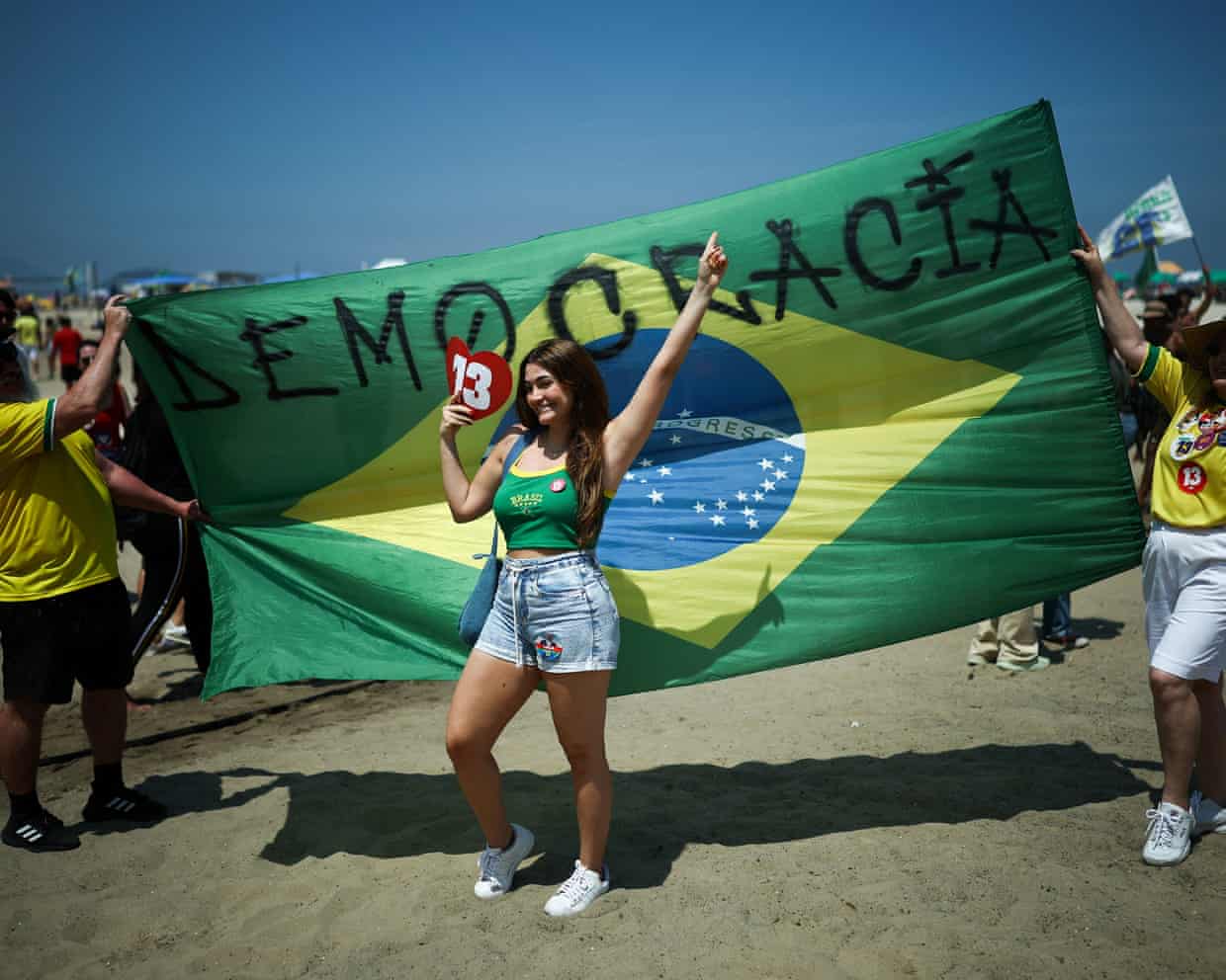 A smiling woman poses before a huge Brazilian flag reading "Democracia" at a Lula rally.