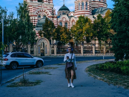 A woman wearing headphones and looking at her phone walks along a road. Behind her is a grand, ornate building