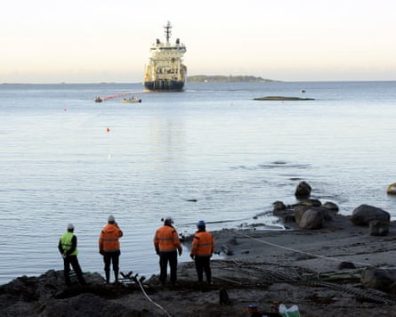 Workers in hi-vis on the shoreline look out towards a large ship in the sea. Cables are trailing from the shoreline into the water.