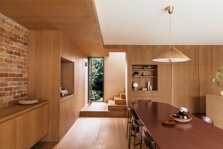 A predominantly wooden house kitchen/living room with a long wooden table, one brick wall, wooden accents and cabinets, wooden floor looking at wooden stairs
