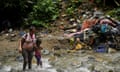 A Venezuelan mother and daughter walk though a river in the jungle. The banks are piled high with discarded clothes and tents.