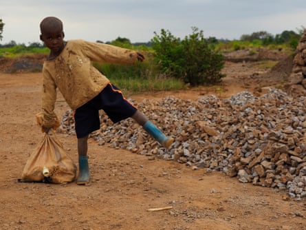 A small boy in a tattered shirt, shorts and rubber boots and holding a sack balances on one leg next to a pile of rocks 