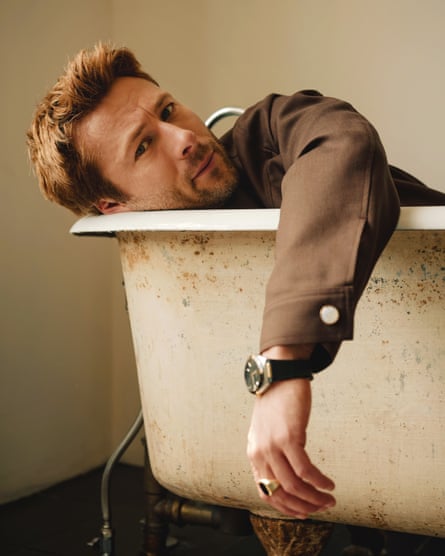 Portrait of Glen Powell wearing a brown jacket and a big watch lying in a rusty bath with his left arm draped over the edge