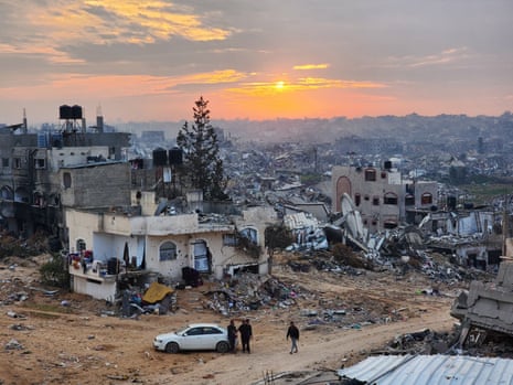 Four people stand near a car, with every visible building destroyed.