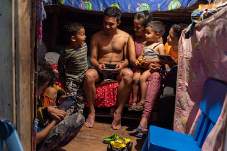 A couple and four children sit together in a small room with wooden walls and material hung up to cover belongings