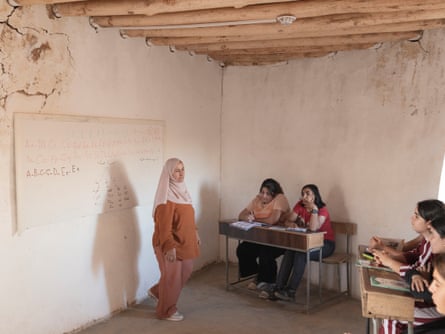 A woman wearing a headscarf stands in front of a whiteboard in a small room. Children sit at desks around the walls