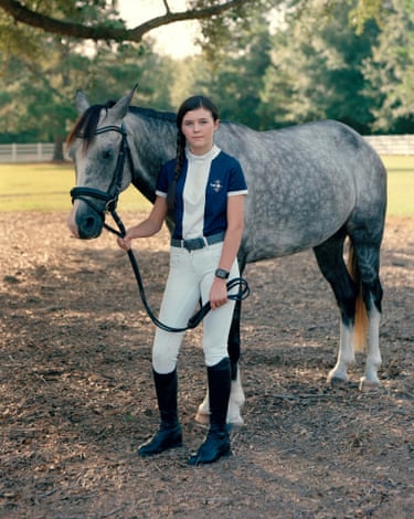 girl in riding gear looks into camera as she stands holding the reins of a horse