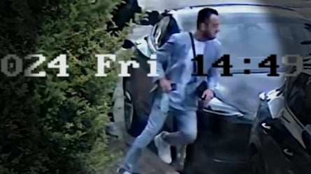 A still taking from CCTV showing a man in a light blue suit starting to run around the front of a parked car