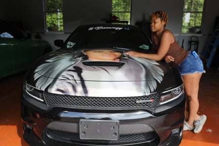 woman leans on car with image on hood of breonna taylor with angel wings. On the top of the windshield, it says #breeway