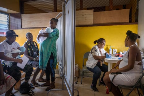A pregnant woman talks to a woman in a white jacket sitting at a table. A screen separates them from another group of people, two of whom are holding babies