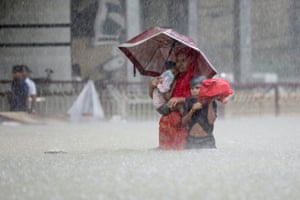 A woman wades through waist-high water in pouring rain while holding a small boy. An older boy walks beside her