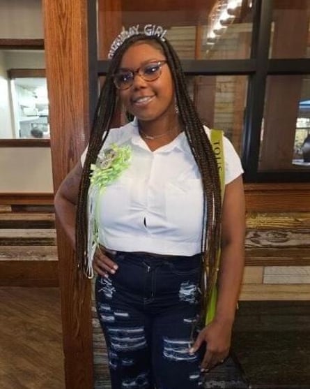 A young-looking Black woman, wearing a tiara that says “Birthday Girl” and a green ribbon corsage on her right chest, with braids past her hips, stands in white blouse, jeans and glasses, posing with one hand on hip, smiling.