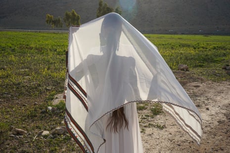 A woman in a white dress stands in a field holding a billowing piece of material over her head and shoulders.