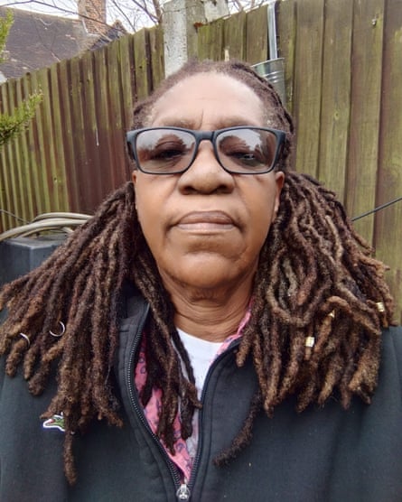 a closeup portrait shows her standing in front of a garden fence, looking directly at the camera; she wears black-framed glasses and a black zip-fronted top, and has neat shoulder-length locks.
