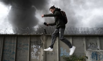 A young person jumps while black smoke rises in the background