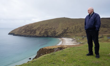 An old Irishman stands on a cliff looking out to sea with a beach curving behind him 