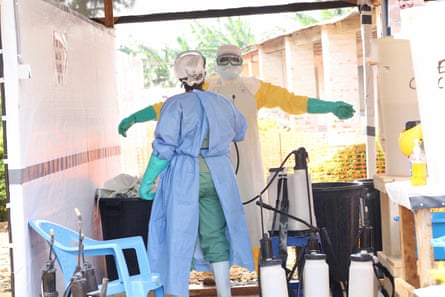 Two medical workers in masks, goggles, long gloves and coveralls stand in a makeshift-looking unit with canisters of disinfectant. One is spraying the other.