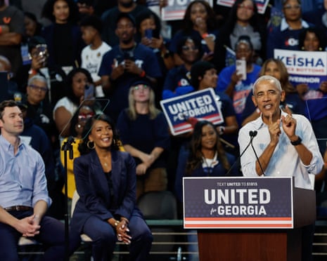 Obama at lectern, smiling and talking, with man and woman seated behind him, smiling.