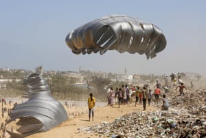 People surround large silver parachutes on a dusty expanse of land. There are mounds of rubbish and rubble nearby