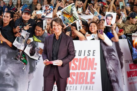 man in suit smiles on red carpet as fans stand behind him holding posters