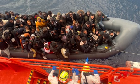 Un bote repleto de personas es remolcado hacia un barco naranja