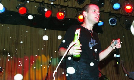 Mike Skinner of the Streets, in a Nike T-shirt, with very short hair and wearing a necklace, holds a microphone in one hand and a bottle of beer in the other
