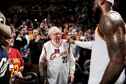 In basketball arena, old white man in Cavaliers jersey greets very tall Black man, also in Cavaliers jersey.