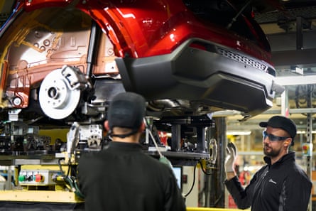 Two Nissan employees stand underneath a part-built red vehicle on the production line. It is still without its wheels.