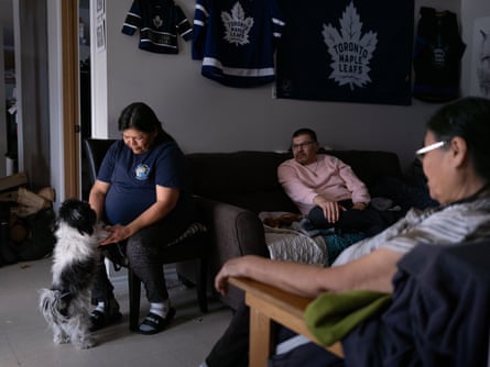 Donald Meeseetawageesic sits with his sister Karen and their mother in their living room; Karen is petting a black and white dog, and there are Toronto Maple Leafs ice-hockey shirts and a flag on the wall behind them.
