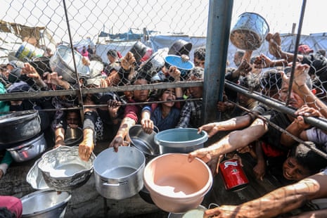 Palestinians stretch their arms through a metal fence with empty pots and pans, trying to receive food from a charity distribution point in Khan Younis, Gaza.