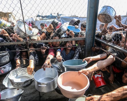 People holding bowls stretch their arms through an opening in a fence for food