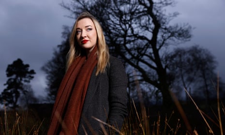 Iona Fyfe, photographed at Harperrig, West Lothian, standing outdoors in wintry weather in front of tree. She has long blond hair and is wearing a black coat, dark red woollen scarf and bright red lipstick