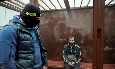 A man sits on a bench flanked by a member of security services in a balaclava