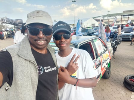 A man and a boy wearing baseball caps and sunglasses take a selfie in a car park