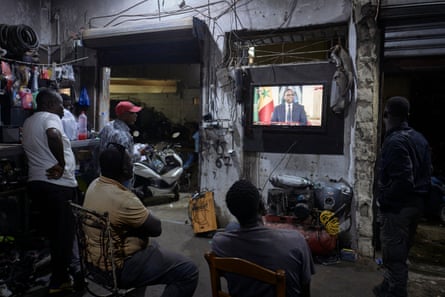 Five men in a workshop watch a man sitting by a flag give an address on TV