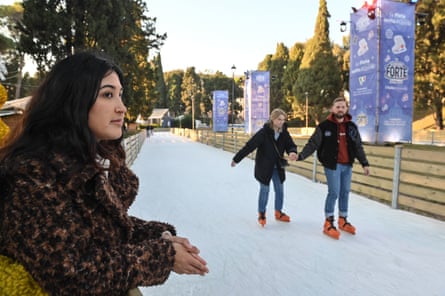 Leila stands at a wooden fence at the edge of a strip of ice, on which two people are skating; she is young with long dark hair, and wears a fake-fur jacket.