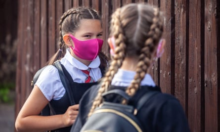 Two girls wearing school uniforms and face masks stand next to a wooden fence
