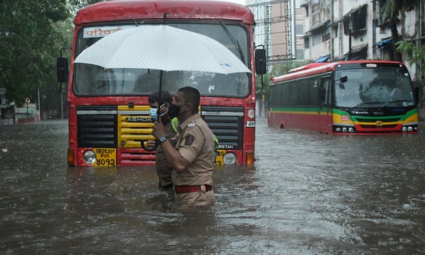 Man in uniform helps another man across a flooded street while water rises to waist height and covers the wheels of a bus