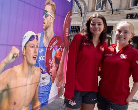 Team England athletes Gabrielle Idle-Beavers and Evie Lambert smile beside promotional banners featuring athletes
