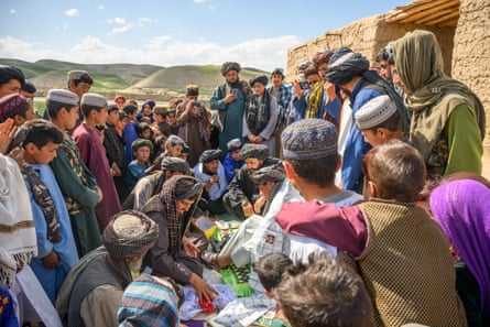 A crowd of men and boys gather around a seated boy, who is being helped to dress, while a man speaks into a microphone