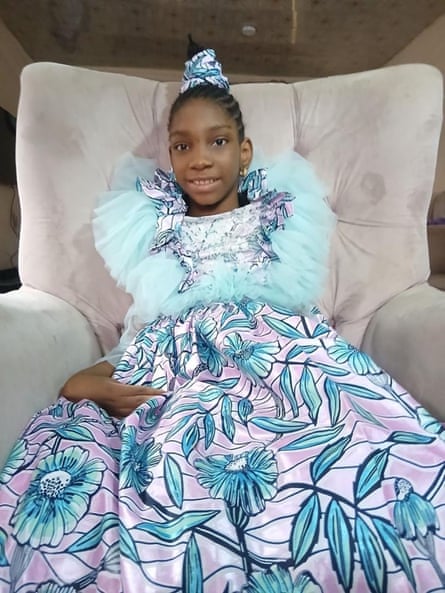 A young woman in a purple and turquoise dress with frills around the collar and matching hairband, sitting on a soft armchair