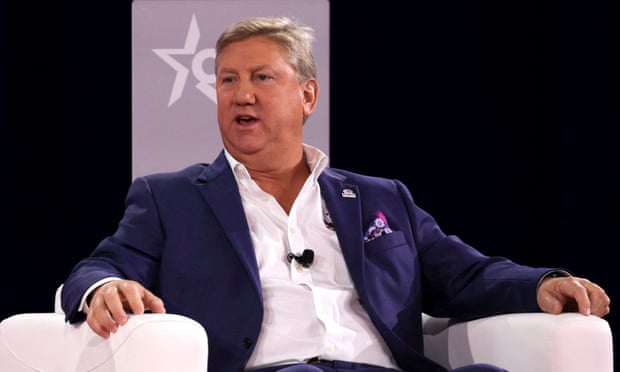 A man dressed in a blue suit speaks while seated on a white chair, with both hands resting on armrests on either side