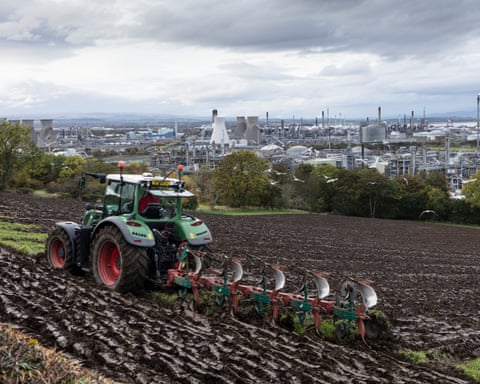 Um trabalhador rural ara os campos com vista para a refinaria e planta petroquímica de Grangemouth, na Escócia.