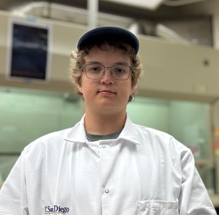 a young man in a while lab coat and baseball cap stands for a photo