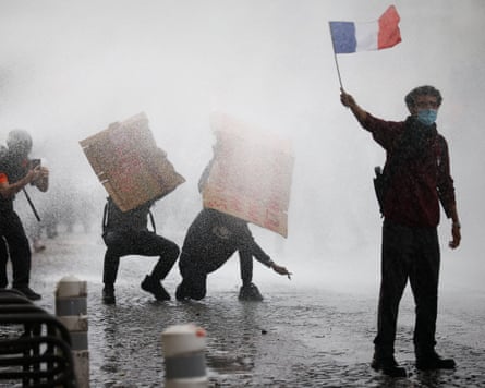 A masked protester holds a French flag as two others behind him use their placards to shield themselves from water canon fire