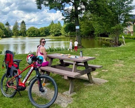 Children in background, bike and picnic table with woman sitting on bench in foreground. Large pond