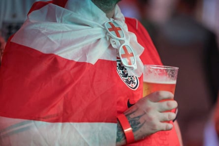 A man, pictured from the shoulders down, holds a pint while wrapped in an England flag, with England sunglasses hanging from his collar