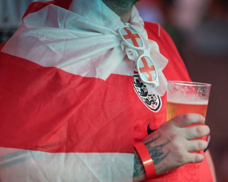 A person in an England flag cape and sunglasses holds a plastic cup of beer